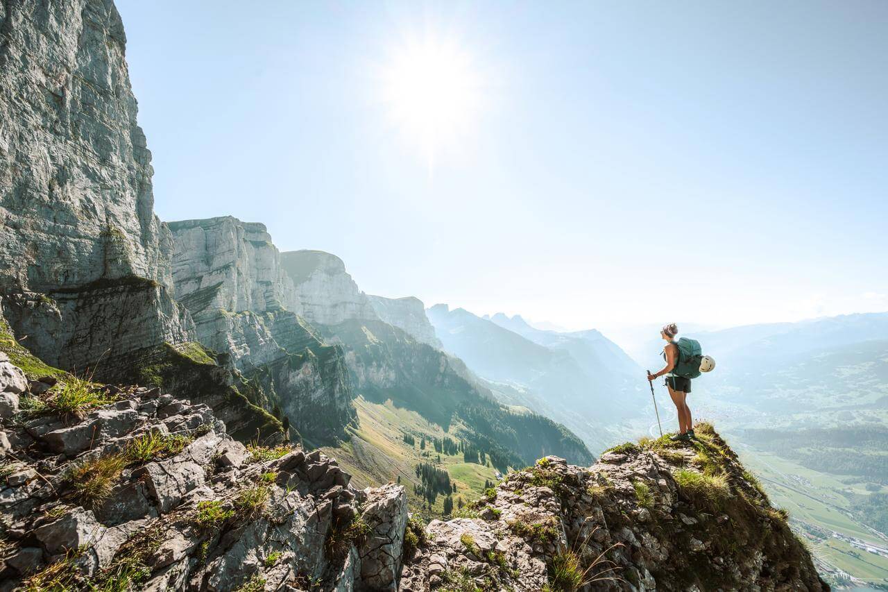 Eine Person steht auf einem felsigen Berggipfel und blickt in die Ferne. Der Himmel ist klar und die Sonne scheint hell. Im Hintergrund sind steile Bergwnde und ein weites Tal sichtbar.