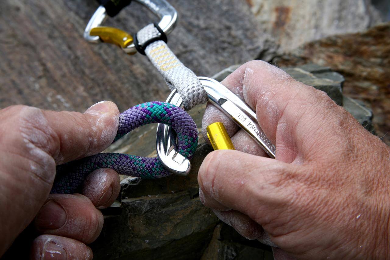Nahaufnahme von zwei Hnden, die Kletterausrstung bedienen. Eine Hand hlt ein buntes Kletterseil, whrend die andere einen Karabiner sichert. Die Ausrstung scheint an einer Kletterwand im Freien verwendet zu werden.