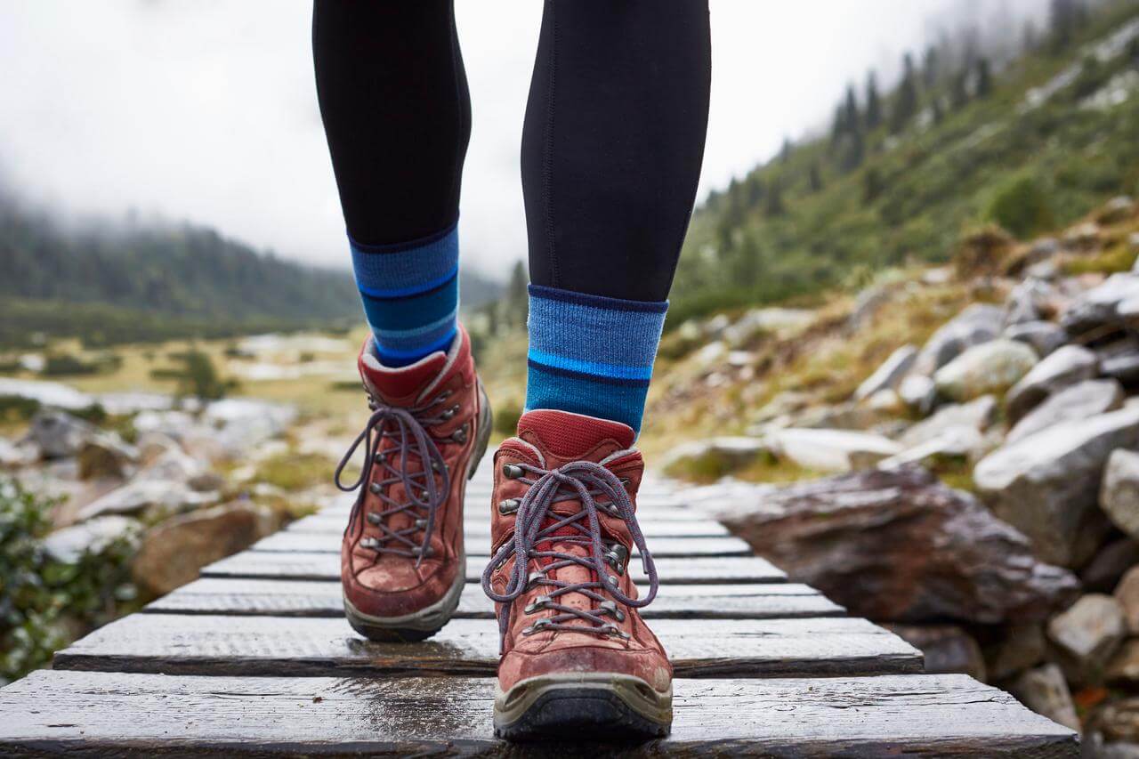 Nahaufnahme von einem Paar wandernden Fen in roten Wanderschuhen und blauen Socken, die auf einem Holzsteg in einer bergigen Landschaft spazieren. Im Hintergrund sind unscharf Grnflchen und Felsen zu erkennen, die von einer wolkigen, nebligen Atmosphre umgeben sind.
