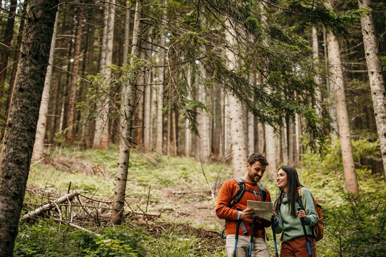 Ein Mann und eine Frau wandern im Wald und betrachten eine Karte. Beide tragen Ruckscke und Outdoor-Kleidung. Die Umgebung ist voller Bume und grnem Laub.