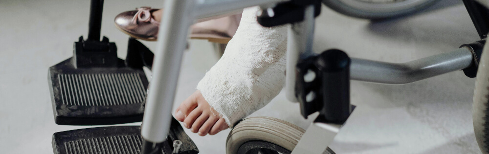 Nahaufnahme eines Fues in einem Gipsverband, der auf der Fusttze eines Rollstuhls ruht. Der andere Fu trgt einen rosa Schuh. Der Rollstuhl hat schwarze Fusttzen und silberne Rahmen.